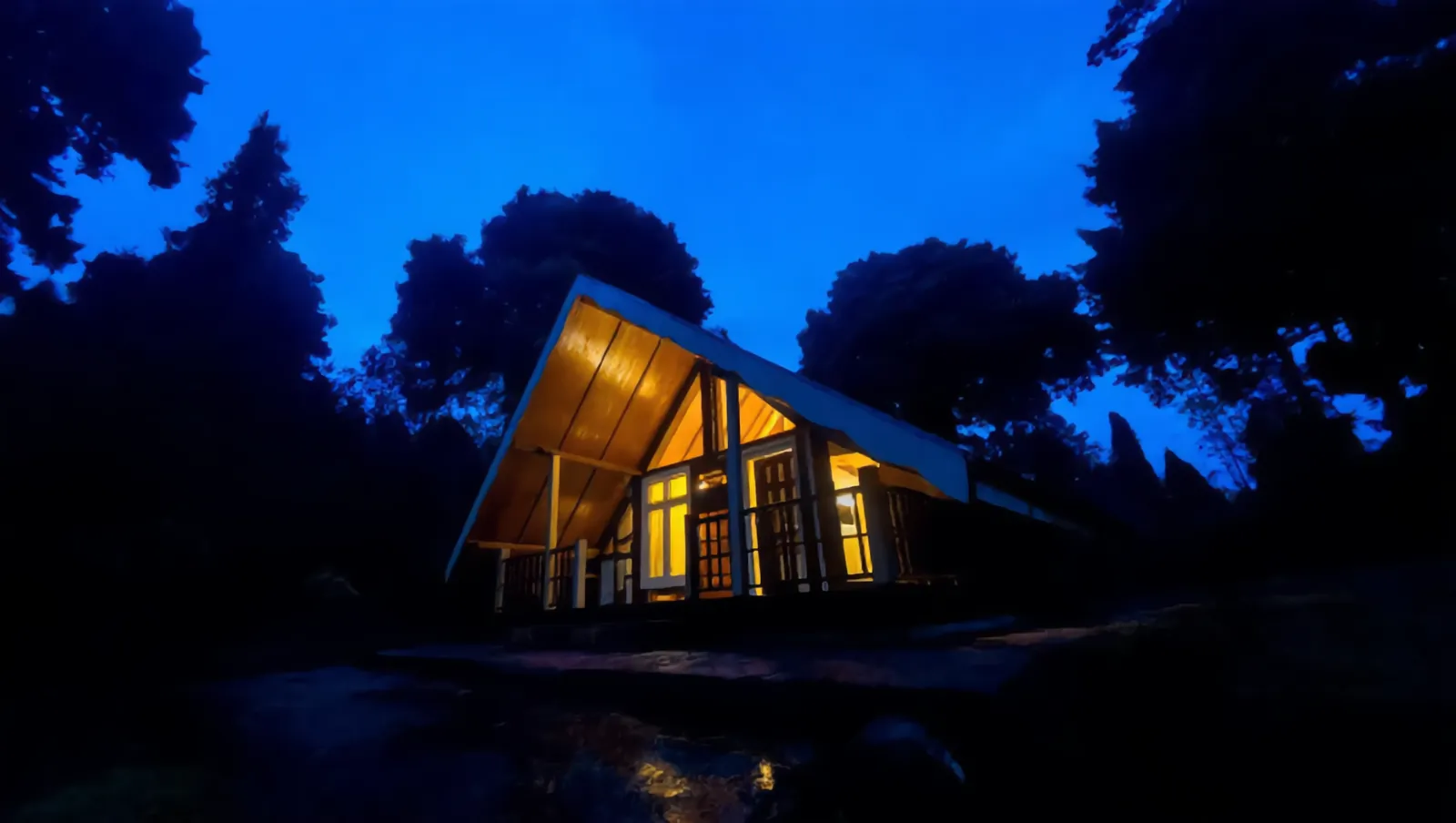 A warmly lit A-frame cabin surrounded by dark trees beneath a deep blue evening sky.